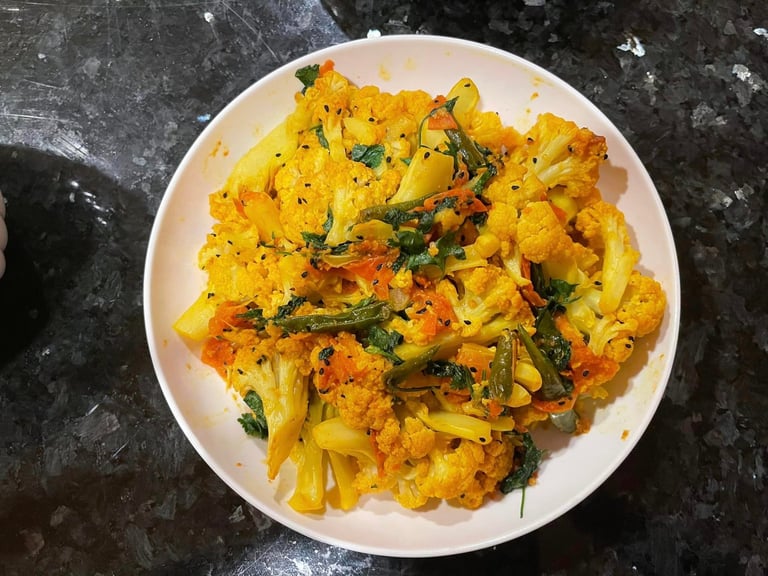 This is a popular Indian vegetarian dish made with cauliflower and potatoes cooked in a flavorful curry sauce. The dish appears to have a vibrant yellow color, likely from the use of turmeric powder, a common spice in Indian cuisine. The presence of green chilies and cilantro suggests a spicy and aromatic flavor profile.
