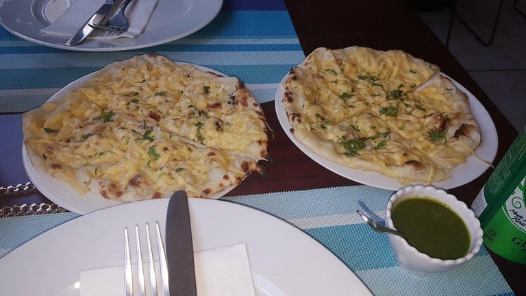 Two herb-topped flatbreads on white plates with a place setting and small bowl of green sauce on a blue checkered tablecloth