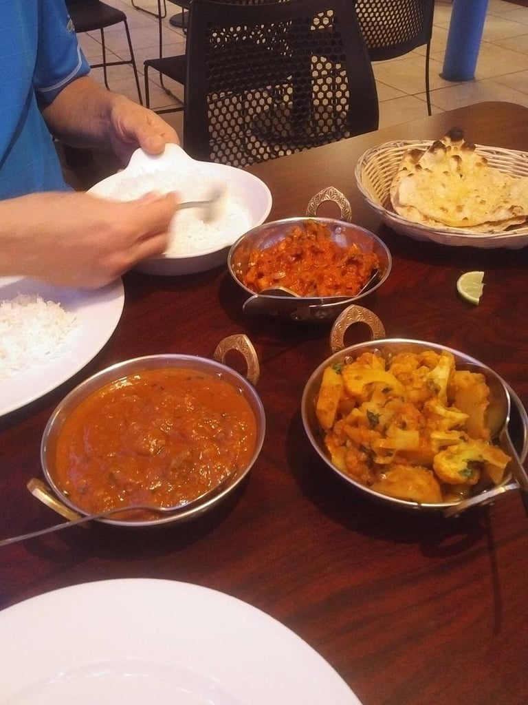 Persons hand holding a white plate at a table with multiple Indian curry dishes in metal bowls and rice on plates