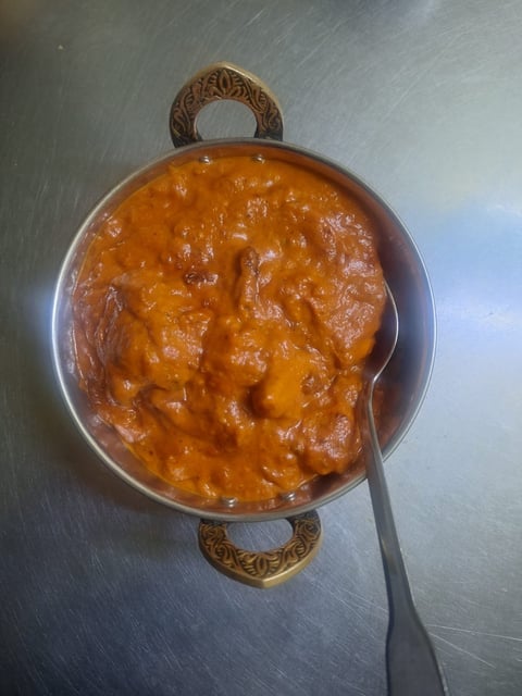 A pot of orange curry or stew with meat and vegetables, topped with an ornate brown handle, on a metal surface